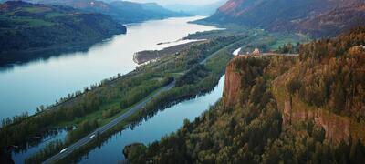 The Columbia River winds between tall reddish brown clifs and green forests.