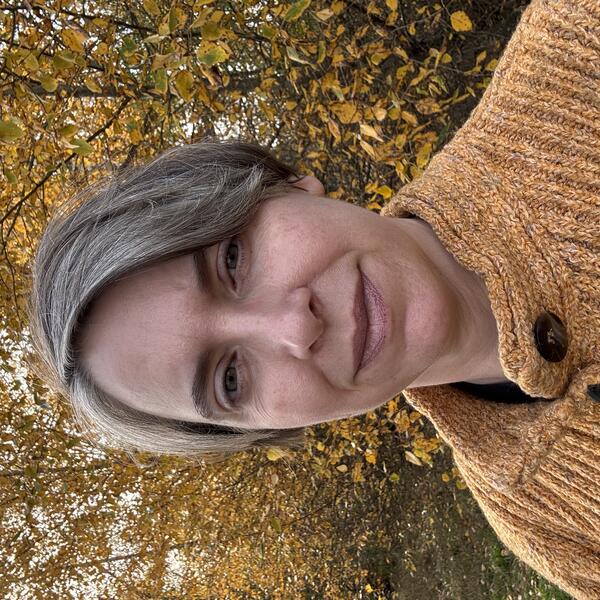  a woman with short hair in an orange sweater in front of trees with yellow leaves